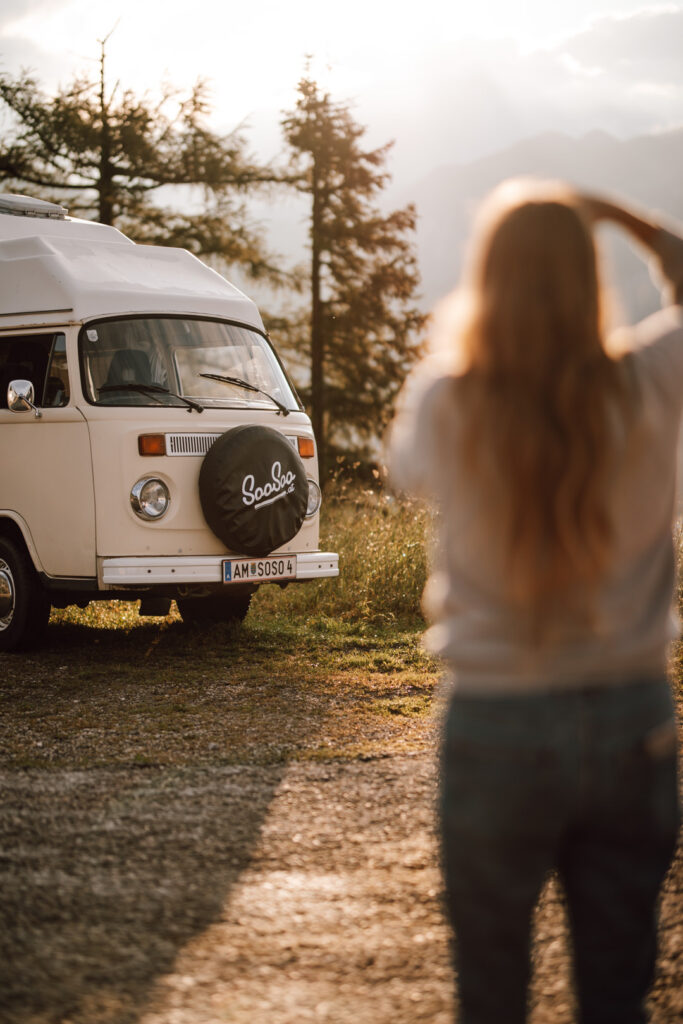 Frau mit langen Haaren fotografiert einen VW Bulli Bus bei Sonnenaufgang am Berg